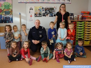Zdjęcie przedstawia zdjęcie grupowe policjanta, przedszkolanki i dzieci w pomieszczeniu