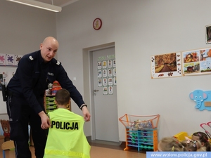 Zdjęcie przedstawia umundurowanego policjanta zakładającego kamizelkę odblaskową z napisem policja chłopcu w pomieszczeniu