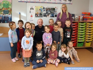 Zdjęcie przedstawia zdjęcie grupowe policjanta, przedszkolanki i dzieci w pomieszczeniu