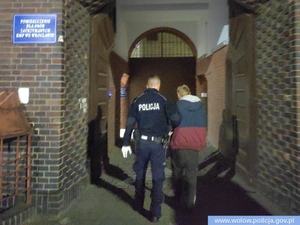 Policjant w mundurze prowadzi inną osobę w kolorowej kurtce przez bramę z cegły, z boku tabliczka z napisem Pomieszczenie Dla Osób Zatrzymanych KWP we Wrocławiu